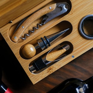 May include: A wooden box containing a set of wine tools, including a corkscrew, a bottle stopper, and a foil cutter. The box is made of light brown wood and has a natural finish.