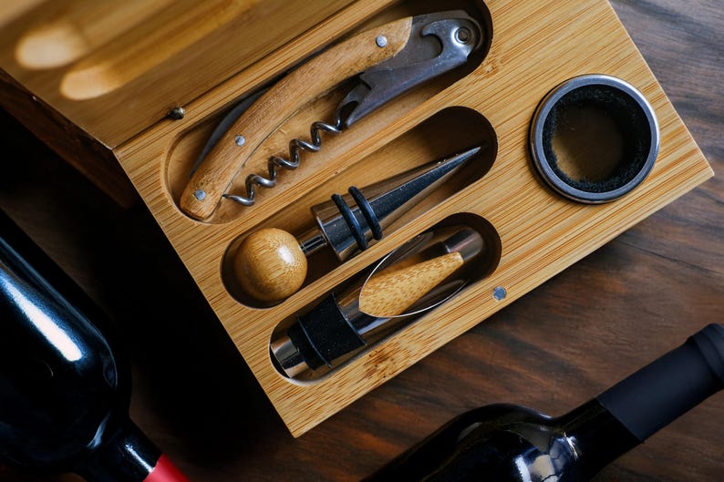 May include: A wooden box containing a set of wine tools, including a corkscrew, a bottle stopper, and a foil cutter. The box is made of light brown wood and has a natural finish.