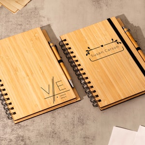May include: Two bamboo notebooks with black spiral bindings and matching pens. One notebook has the initials "V/E" with the text "VANDY & ERNIE" below. The other notebook has the text "Susan Carson" in a rectangular frame with a floral design.