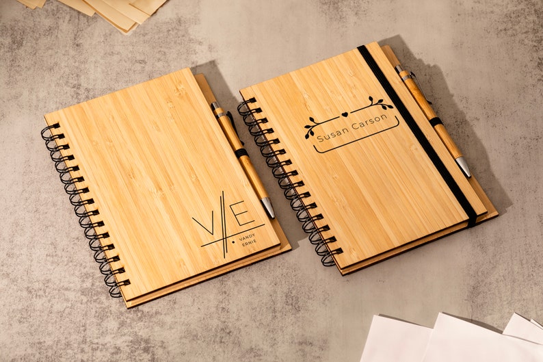 May include: Two bamboo notebooks with black spiral bindings and matching pens. One notebook has the initials "V/E" with the text "VANDY & ERNIE" below. The other notebook has the text "Susan Carson" in a rectangular frame with a floral design.