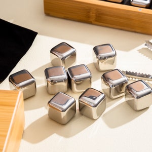 May include: A set of stainless steel ice cubes in a white background. The ice cubes are arranged in a triangular shape. There is a small black pouch and a wooden box in the background.