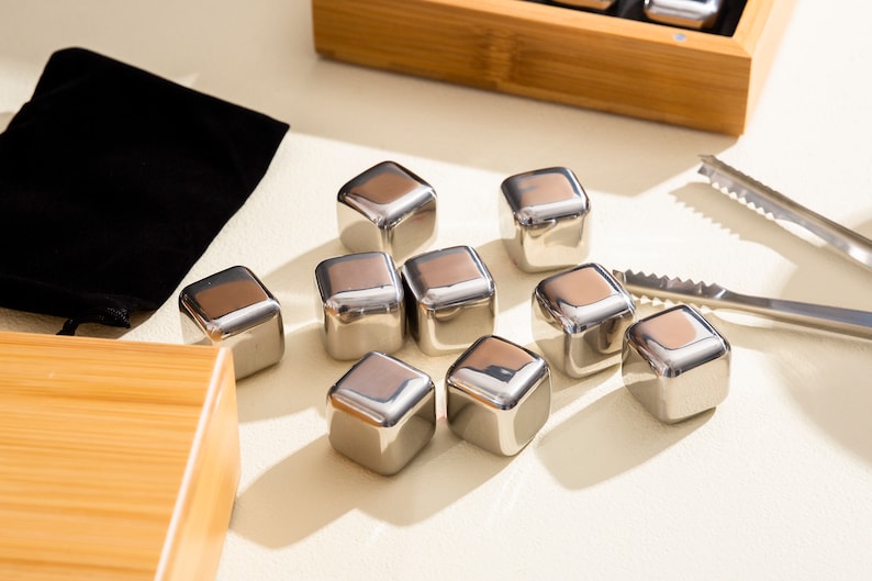 May include: A set of stainless steel ice cubes in a white background. The ice cubes are arranged in a triangular shape. There is a small black pouch and a wooden box in the background.