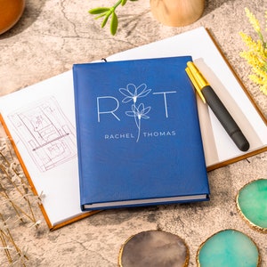 May include: A blue notebook with the initials "R&T" and the names "Rachel Thomas" printed in white. The notebook is closed and lying on a table with a pen and other decorative items.
