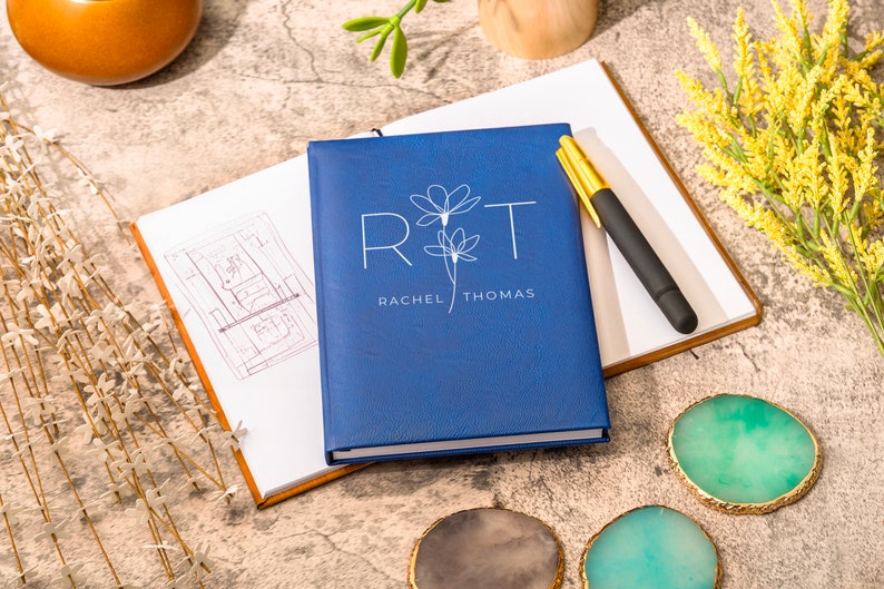 May include: A blue notebook with the initials "R&T" and the names "Rachel Thomas" printed in white. The notebook is closed and lying on a table with a pen and other decorative items.