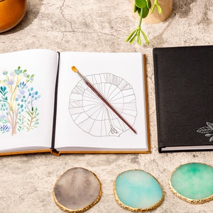 May include: A black leather journal with a white floral design and the name "Olivia" on the cover. The journal is open to a blank page with a paintbrush on top. The journal is surrounded by three agate coasters.