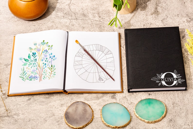 May include: A black leather journal with a white floral design and the name "Olivia" on the cover. The journal is open to a blank page with a paintbrush on top. The journal is surrounded by three agate coasters.