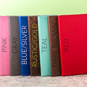 May include: A collection of colorful binders in black/silver, pink, gray, blue/silver, rustic/gold, teal, dark brown, and red. Each binder has a vertical text label with its color name. The binders are arranged in a row.