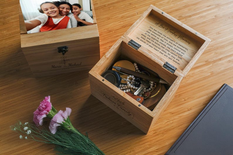 May include: A wooden box with a hinged lid, engraved with the words "A Box of Family Memories". The box is open, revealing a small collection of trinkets and a photo of three people. The box is labeled "Our Family" on the front.