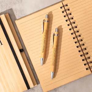 May include: Two bamboo pens with personalized names, "Teresa Canfield PhD" and "Ashley Sanford", are lying on a lined notebook with a bamboo cover. The notebook has a black elastic band and a black spiral binding. The notebook cover has the initials "TCM" engraved on it.