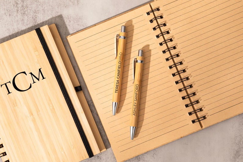 May include: Two bamboo pens with personalized names, "Teresa Canfield PhD" and "Ashley Sanford", are lying on a lined notebook with a bamboo cover. The notebook has a black elastic band and a black spiral binding. The notebook cover has the initials "TCM" engraved on it.