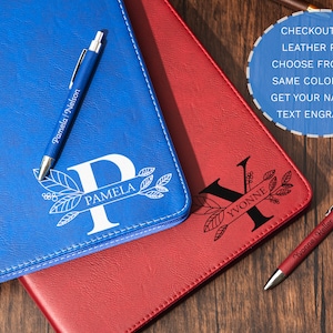 May include: Blue and red leather journals with personalized initials and names, accompanied by matching pens. The blue journal features the initial "P" and the name "Pamela", while the red journal displays "Yvonne". The pens are also engraved.