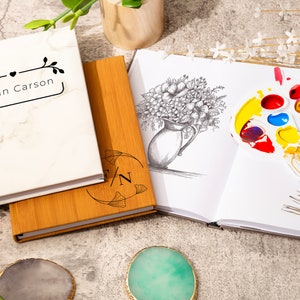 May include: Two blank journals, one with a marble cover and the text "Susan Carson" and the other with a wooden cover and the text "S/N". A sketchbook with a pencil drawing of a bouquet of flowers in a vase and a paint palette with colorful paint.