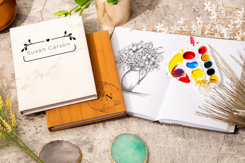 May include: Two blank journals, one with a marble cover and the text "Susan Carson" and the other with a wooden cover and the text "S/N". A sketchbook with a pencil drawing of a bouquet of flowers in a vase and a paint palette with colorful paint.