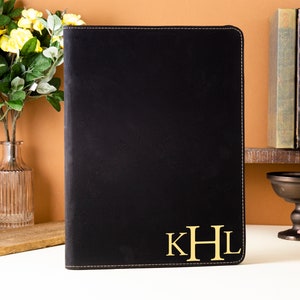 May include: Black leather portfolio with gold-colored initials "KHL" on the lower right corner. The portfolio has a stitched border. The background includes a vase of yellow flowers, books, and decorative items.