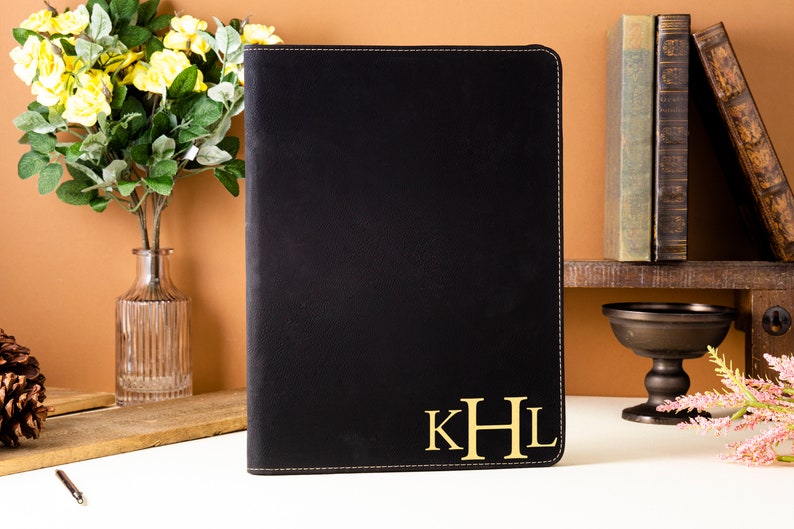 May include: Black leather portfolio with gold-colored initials "KHL" on the lower right corner. The portfolio has a stitched border. The background includes a vase of yellow flowers, books, and decorative items.