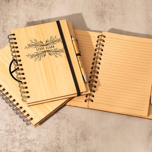 May include: Two bamboo notebooks with spiral binding and a pen. One notebook has the name "Steve Clark" engraved on the cover. The other notebook has a peace sign engraved on the cover.