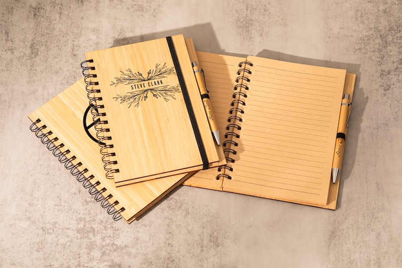 May include: Two bamboo notebooks with spiral binding and a pen. One notebook has the name "Steve Clark" engraved on the cover. The other notebook has a peace sign engraved on the cover.