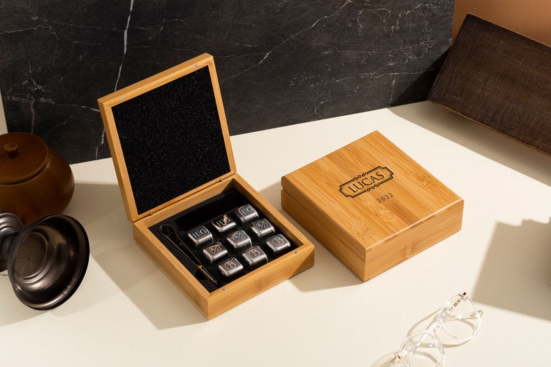 May include: A wooden box with a personalized inscription "Lucas 2022" on the lid. The box is open, revealing a set of nine silver whiskey stones and a pair of tongs.