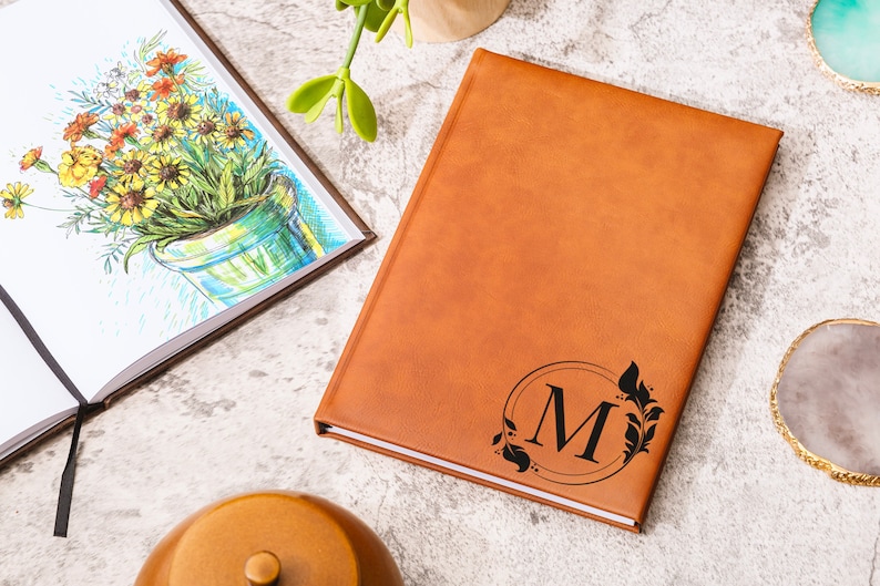 May include: A brown leather journal with a black monogram of the letter "M" surrounded by a circle and floral design.