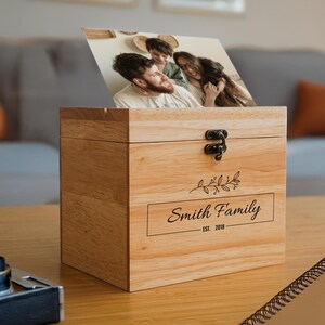 May include: A wooden photo box with a hinged lid and a brass latch. The box has a personalized inscription that reads "Smith Family Est. 2018" and a small floral design. A photo of a family is visible through the open lid.
