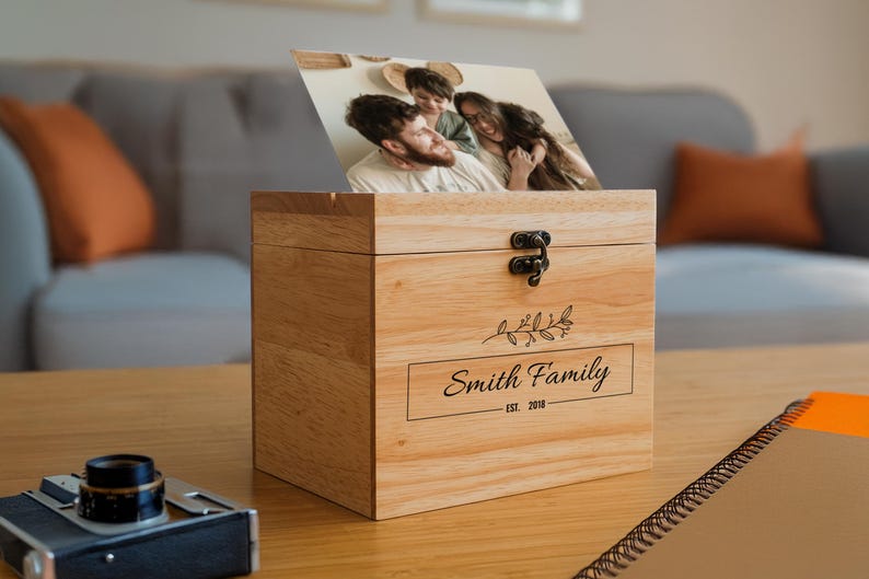 May include: A wooden photo box with a hinged lid and a brass latch. The box has a personalized inscription that reads "Smith Family Est. 2018" and a small floral design. A photo of a family is visible through the open lid.