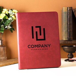 Op de afbeelding: Een rood lederen gebonden dagboek met een zwart logo met de tekst "COMPANY" en "SLOGAN HERE". Het dagboek ligt op een wit oppervlak met een vaas met gele bloemen en een stapel boeken op de achtergrond.