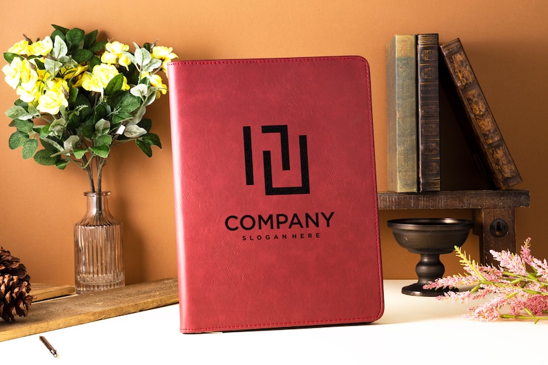Op de afbeelding: Een rood lederen gebonden dagboek met een zwart logo met de tekst "COMPANY" en "SLOGAN HERE". Het dagboek ligt op een wit oppervlak met een vaas met gele bloemen en een stapel boeken op de achtergrond.