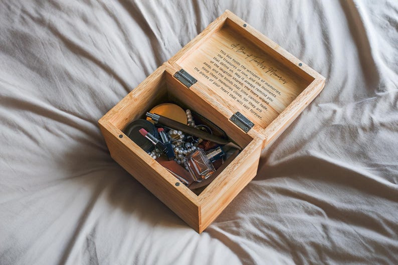 May include: A wooden box with a hinged lid, open to reveal a variety of items, including lipstick, a pen, a small bottle of perfume, and a pearl necklace. The box has a quote inscribed on the inside of the lid: "This box holds the whispers of memories, the treasures of time, and the moments that make us who we are. May these whispers always remind you of the laughter that could not be contained, and the moments that time cannot fade. Keep these memories close, for they are the pieces of your heart that will last forever."