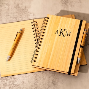 May include: Two bamboo notebooks with black spiral bindings and matching pens. The notebook on top has a black elastic band and the initials "AKM" engraved on the front. Both pens have the name "Aaron Klein" engraved on them.