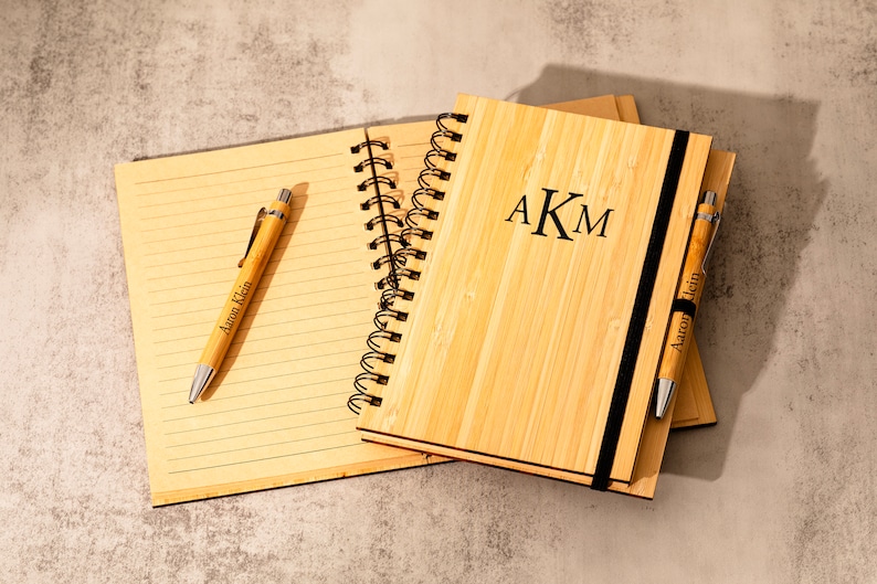 May include: Two bamboo notebooks with black spiral bindings and matching pens. The notebook on top has a black elastic band and the initials "AKM" engraved on the front. Both pens have the name "Aaron Klein" engraved on them.