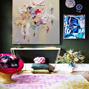 K&ouml;nnte beinhalten: Ein Wohnzimmer-Interieur mit einem farbenfrohen abstrakten Gem&auml;lde, einem modernen Sessel und einem gemusterten Teppich. Das Gem&auml;lde zeigt einen strukturierten, wei&szlig;en Hintergrund mit bunten abstrakten Formen und Figuren. Der Sessel ist leuchtend pink mit einem schwarz-wei&szlig;en flauschigen Kissen. Der Teppich ist ein mehrfarbiger, abstrakter Entwurf in Pink-, Gelb- und Wei&szlig;t&ouml;nen.