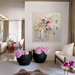 K&ouml;nnte beinhalten: Ein Wohnzimmer-Interieur mit einem gro&szlig;en abstrakten Gem&auml;lde an der Wand, zwei schwarzen Pflanzgef&auml;&szlig;en mit rosa Blumen, einem wei&szlig;en Sofa und einem braunen Kuhfellteppich.