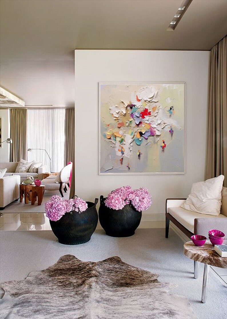 K&ouml;nnte beinhalten: Ein Wohnzimmer-Interieur mit einem gro&szlig;en abstrakten Gem&auml;lde an der Wand, zwei schwarzen Pflanzgef&auml;&szlig;en mit rosa Blumen, einem wei&szlig;en Sofa und einem braunen Kuhfellteppich.