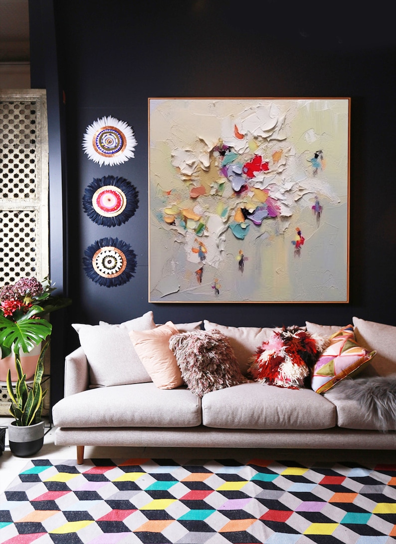 K&ouml;nnte beinhalten: Ein wei&szlig;es Sofa mit bunten Kissen steht vor einer dunkelblauen Wand mit einem gro&szlig;en abstrakten Gem&auml;lde und drei dekorativen Wandbeh&auml;ngen. Ein bunter geometrischer Teppich bedeckt den Boden.