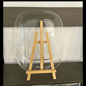 May include: A clear, oval-shaped glass tray is displayed upright on a light-colored wooden easel. The tray features a smooth, transparent surface with rounded edges, providing a simple and functional design for holding various items.