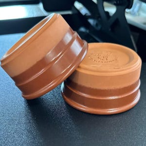 May include: Two round terracotta ramekins featuring a two-tone design with a lighter matte upper section and a darker glazed lower section. The base of the right ramekin displays a stamped circular logo with a fork and knife icon and text.