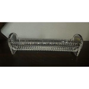 May include: A clear glass elongated tray featuring a distinct vertical ribbed pattern along its entire length. The tray has a semi-cylindrical shape with rounded, elevated ends that serve as supports, creating a structured and transparent appearance.