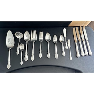 May include: A set of silver-toned flatware including a cake server with a perforated blade, a ladle, various spoons, forks, and knives. The utensils feature a consistent decorative handle design with a rounded end and a central ridge.