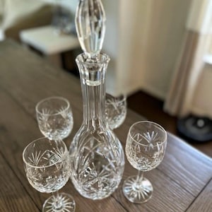 May include: A clear cut glass decanter with a matching stopper and four matching stemmed glasses. The decanter and glasses feature a starburst and cross-hatch pattern. A portion of a ruler is visible in the corner of the frame.