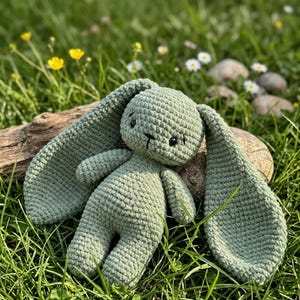 May include: A handmade, sage green crochet bunny with long floppy ears, black eyes, and a stitched mouth. The plush toy is lying on a rock and a piece of wood in a grassy area with wildflowers.