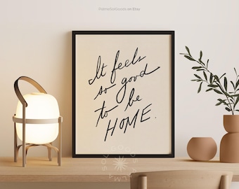 May include: A framed print with the text "It feels so good to be HOME" in black script. A glowing white lamp with a handle sits to the left. A small vase and decorative plant are on the right, all on a wooden shelf.