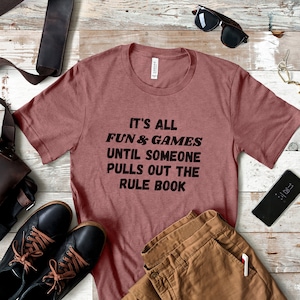 May include: A maroon t-shirt with the text "It's all fun & games until someone pulls out the rule book" printed in black.