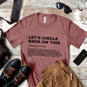 May include: A maroon t-shirt with the text "Let's Circle Back On This" in black. The text is defined as an "office jargon" phrase and describes an overused expression used by management to avoid actually doing any work or responding to questions. The origin of the phrase came from Anita Dayhoff, an overworked employee, who observed pigeons on her lunch break and noticed that they aimlessly walk in circles for no reason.