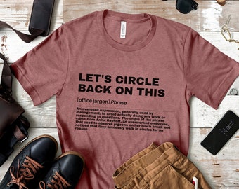 May include: A maroon t-shirt with the text "Let's Circle Back On This" in black. The text is defined as an "office jargon" phrase and describes an overused expression used by management to avoid actually doing any work or responding to questions. The origin of the phrase came from Anita Dayhoff, an overworked employee, who observed pigeons on her lunch break and noticed that they aimlessly walk in circles for no reason.
