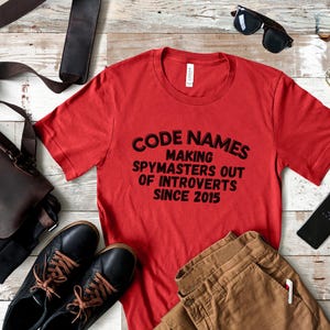 May include: Red t-shirt with black text that reads "Code Names Making Spymasters Out of Introverts Since 2015".