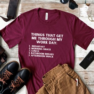May include: A maroon t-shirt with white text that reads "Things that get me through my work day: 1. Breakfast 2. Morning Snack 3. Lunch 4. Bathroom Breaks 5. Afternoon Snack".