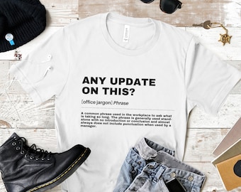 May include: White t-shirt with a black graphic design that reads "ANY UPDATE ON THIS? [office jargon] Phrase" and a definition of the phrase.