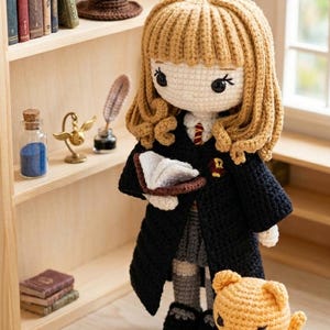 Puede incluir: Muñeca de ganchillo hecha a mano, con cabello rubio, vestida con una túnica negra y sosteniendo un libro. También se ven un pequeño gato naranja y un sombrero puntiagudo marrón. La muñeca está de pie frente a una estantería de madera con libros en miniatura y objetos decorativos.