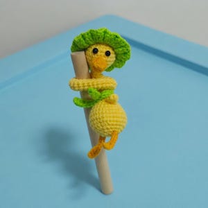 Könnte beinhalten: Ein handgefertigtes Häkelspielzeug in Form eines Entchens in Gelb, Grün und Orange, das auf einem Holzdübel sitzt. Das Entchen hat einen grünen Hut und hält sich mit Armen und Beinen an dem Dübel fest. Der Hintergrund ist eine hellblaue Oberfläche.