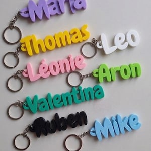 May include: A collection of eight custom name keychains in various colors including purple, yellow, white, pink, green, teal, black, and blue. Each keychain features a metal ring attached to a 3D-printed name: Maria, Thomas, Leo, Léonie, Aron, Valentina, Ruben, and Mike.